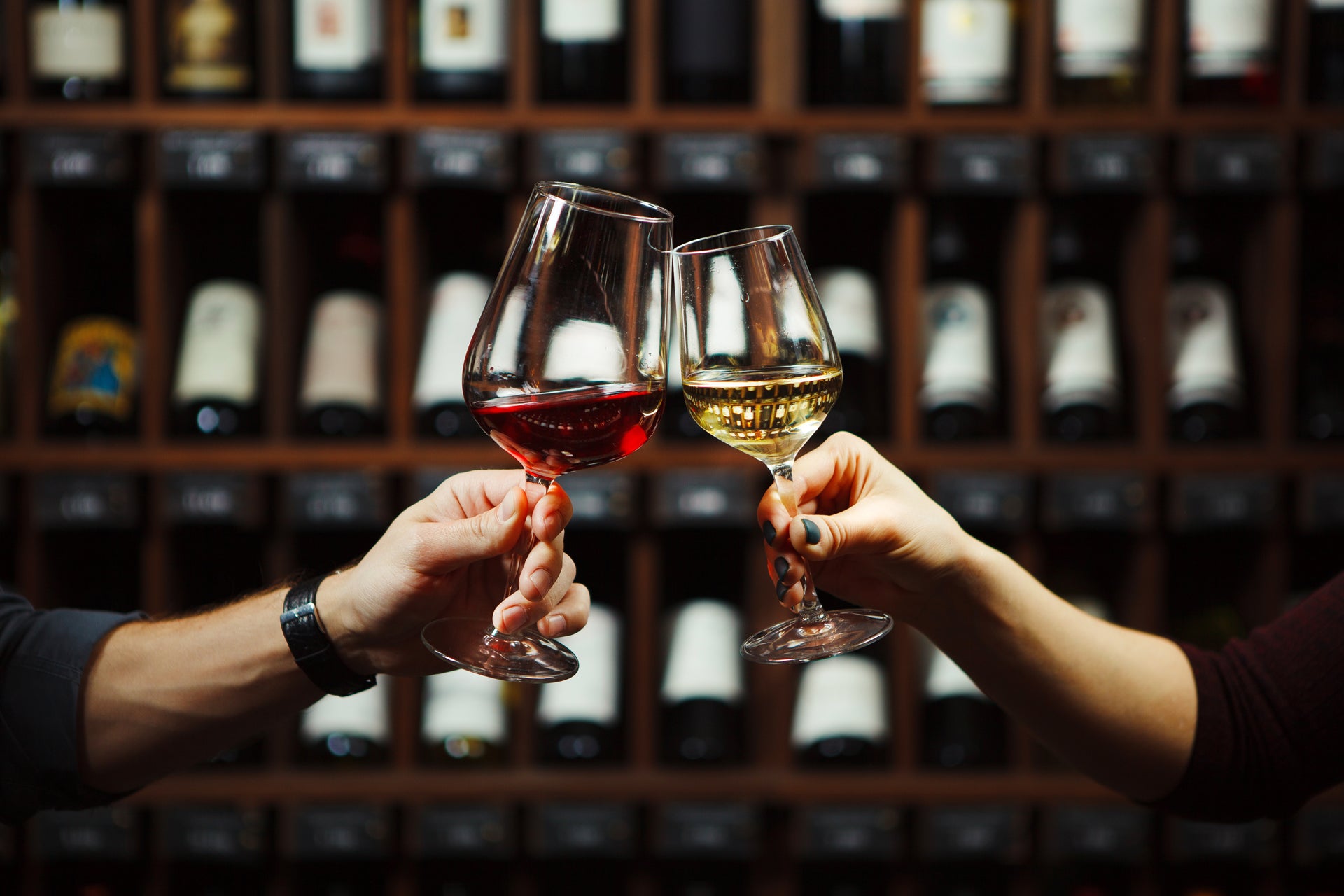 A photograph of a man's left hand holding a glass of red wine touching a glass of white wine held in a woman's right hand in front of shelves of wine bottles